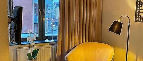 Habitación de hotel con cama, una silla, una mesita con planta, ventana con cortinas y lámpara.