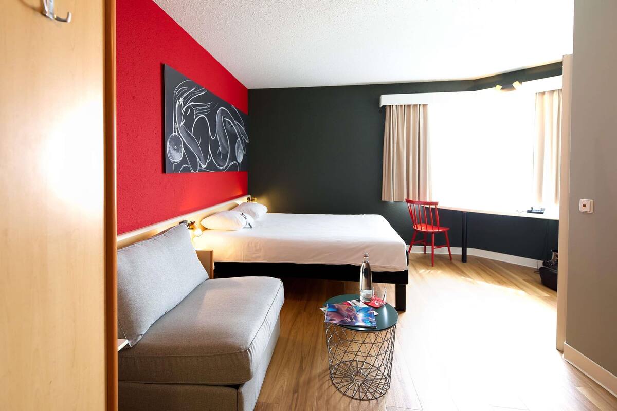 A hotel room with a bed, a sofa, a red chair, a small round table with a magazine, and a large abstract painting on the wall.