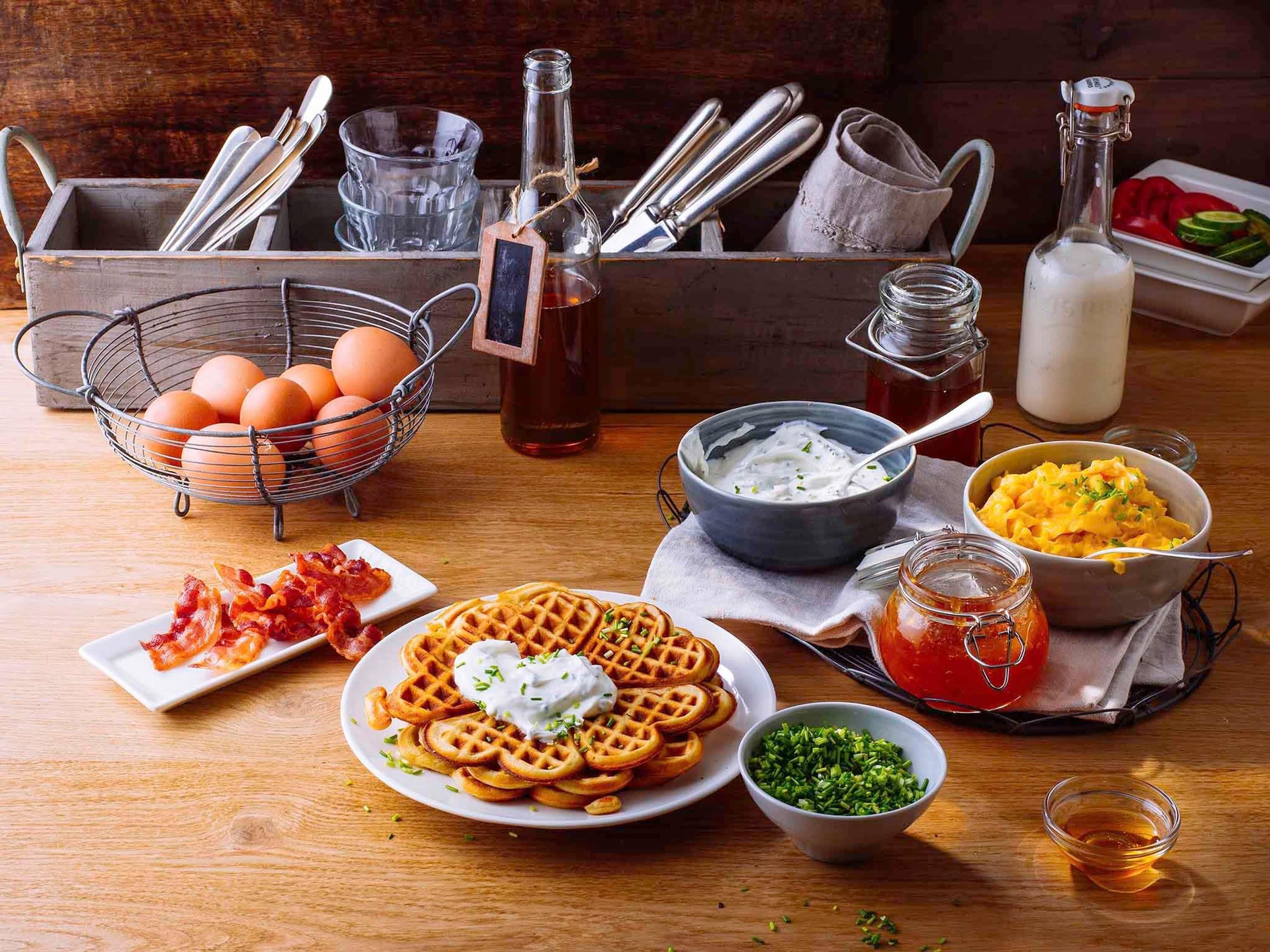 A breakfast table with waffles, eggs, bacon, yogurt, and various condiments.