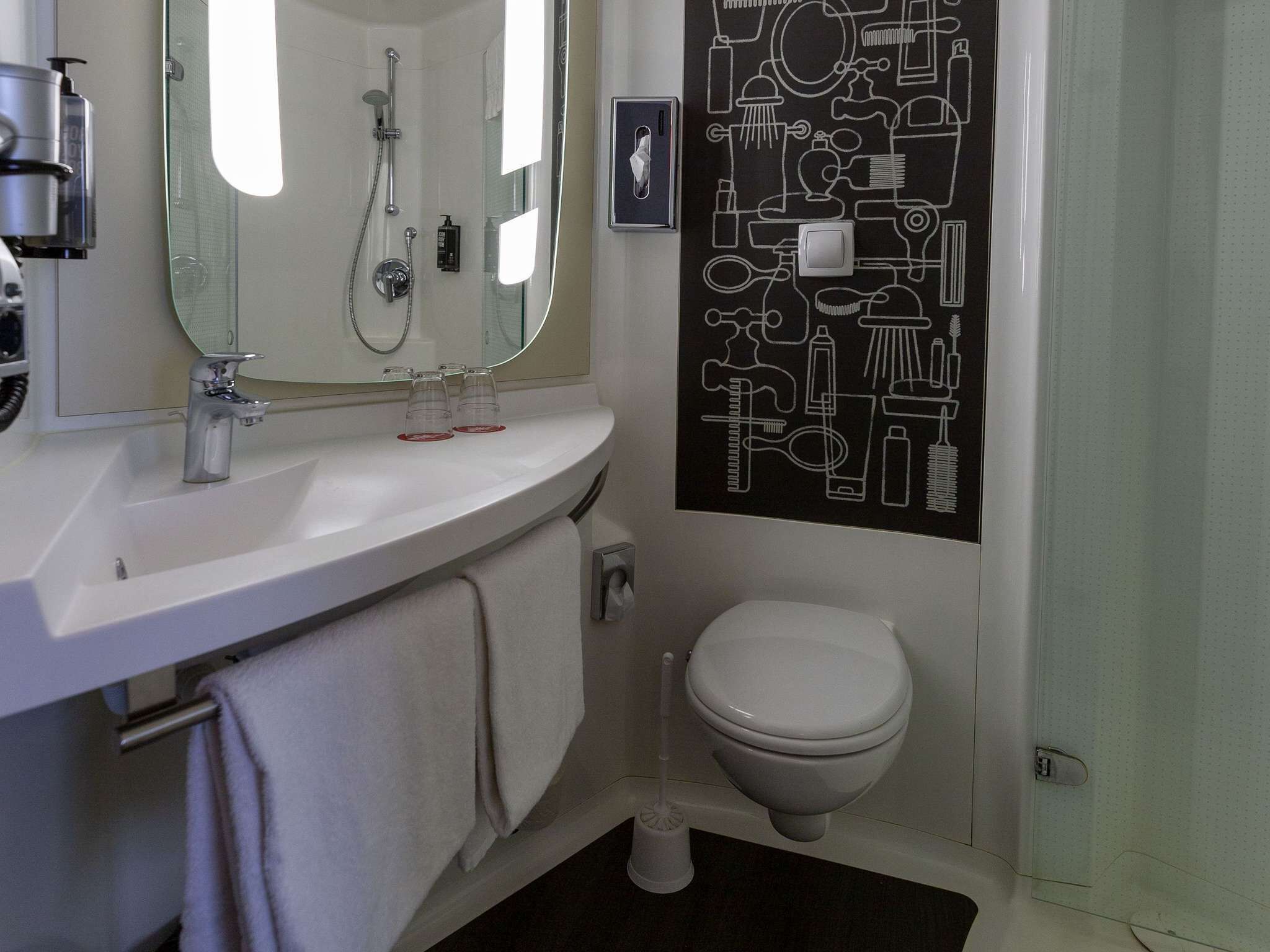 Un baño con lavamanos blanco, espejo, ducha, inodoro y perchero para toallas.
