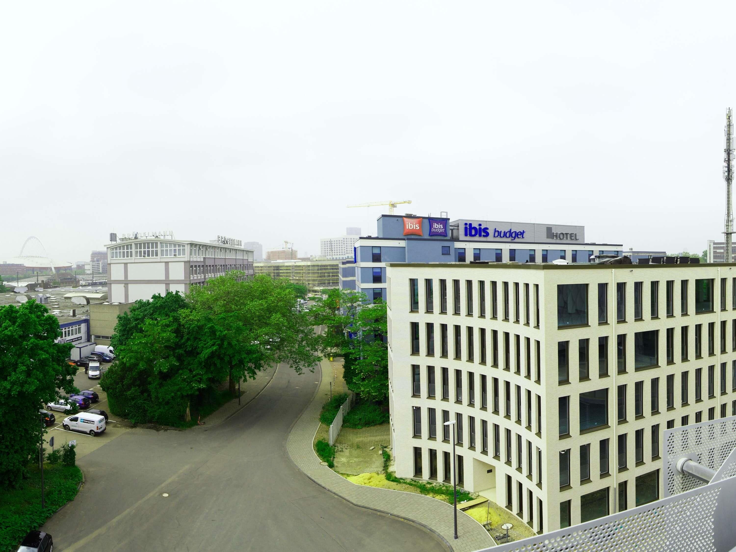 A modern building with 'ibis budget' signage, a parking lot, and surrounding greenery.