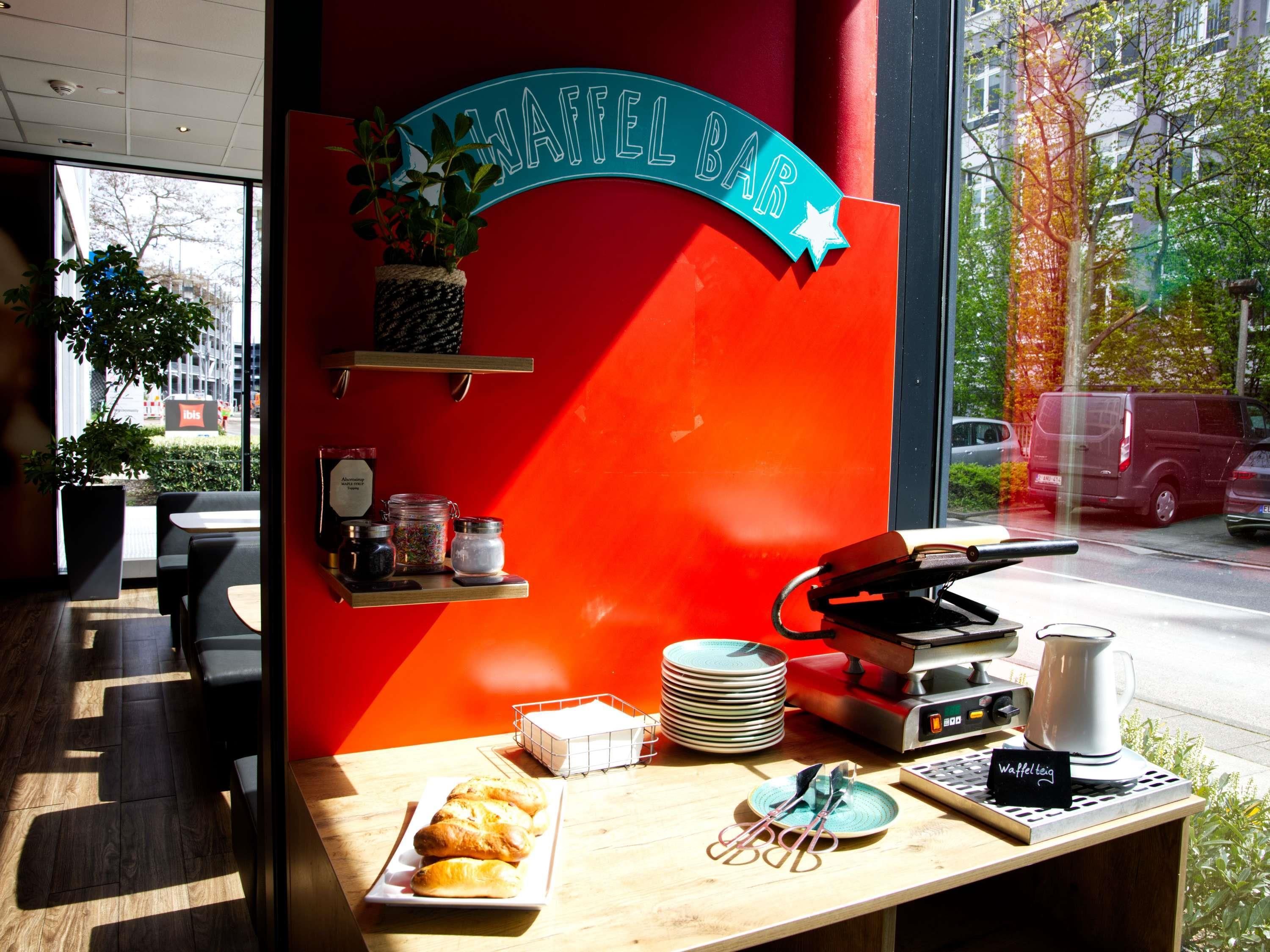 Ein Frühstücksbuffet mit roter Wand, einem Schild mit der Aufschrift "WAFFEL BAR" und verschiedenen Frühstücksangeboten wie Brot und Kaffee.