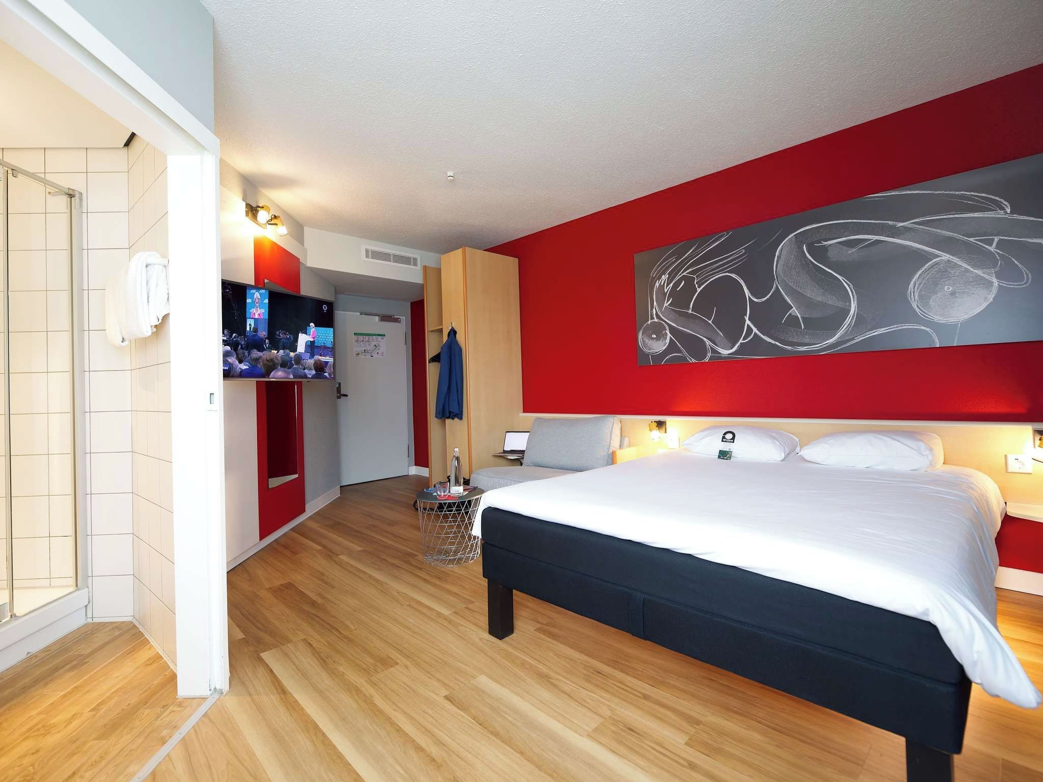 Ein Hotelzimmer mit einem großen Bett, einem Fernseher, einer roten Akzentwand und einem Holzboden.