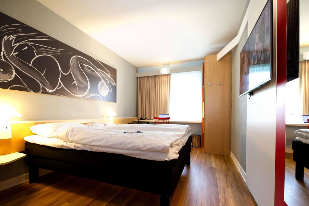 A hotel room with a bed, bedside lamps, a desk with a chair, a TV, and a large abstract painting on the wall.