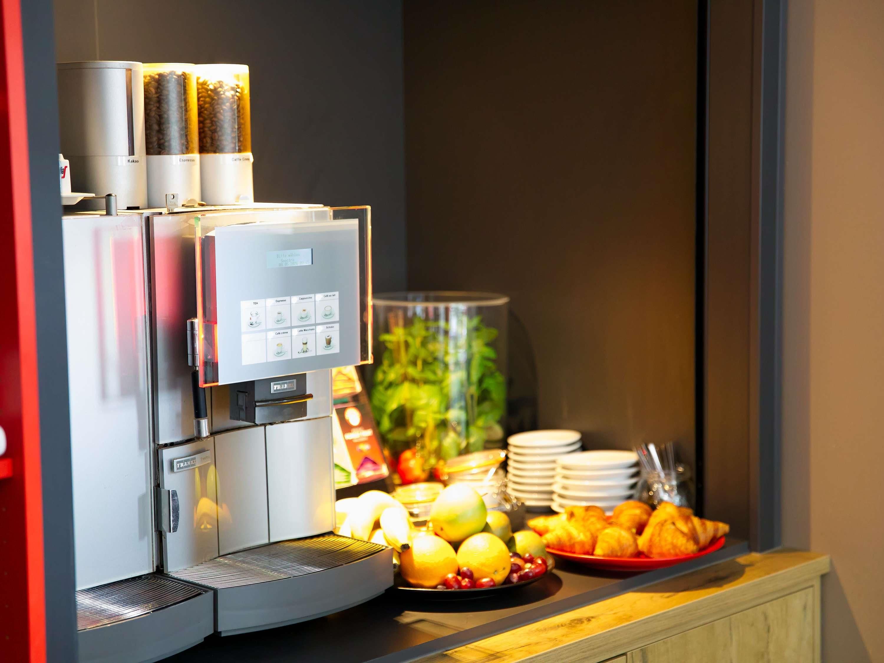 Una máquina de café con varios dispensadores, una selección de frutas y bollería sobre una encimera.