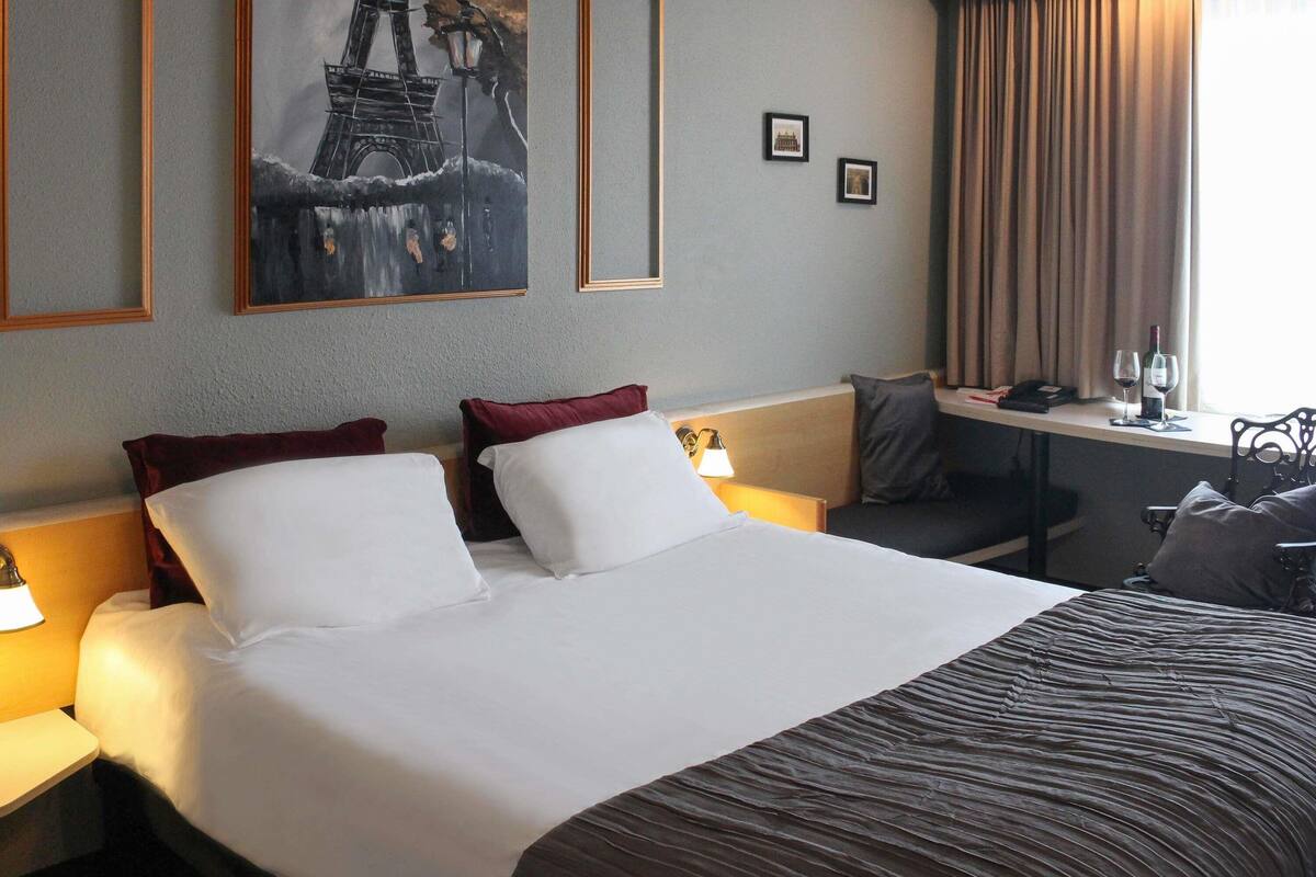 A hotel room with a large bed, a desk with a lamp, a chair, and a window with curtains.