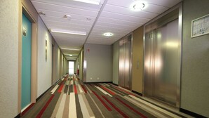 Un long couloir étroit, typique des hôtels, avec un tapis à motifs, des portes sur le côté gauche et des ascenseurs sur le côté droit.
