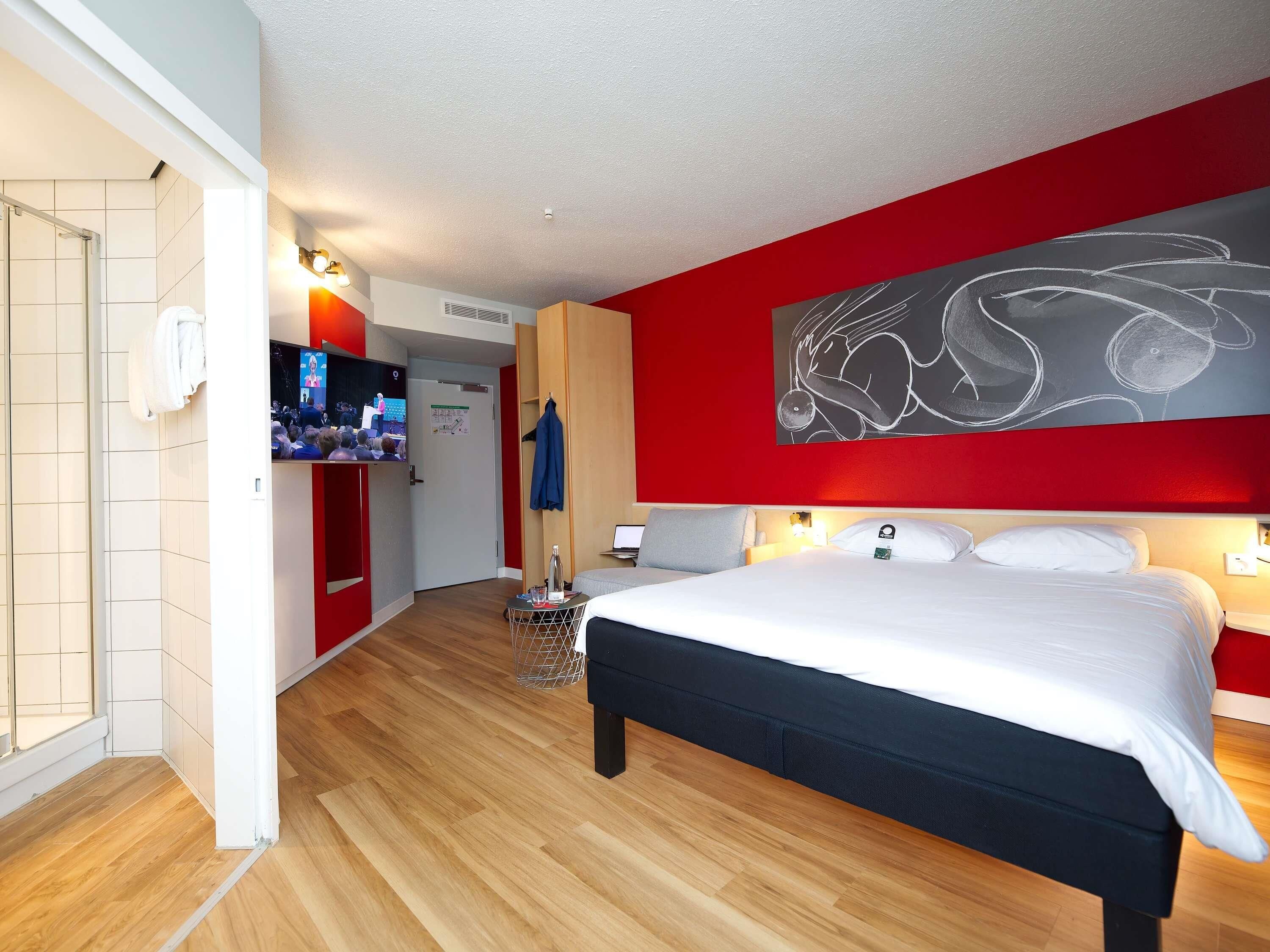 A modern hotel room with a large bed, a television, and a red accent wall.