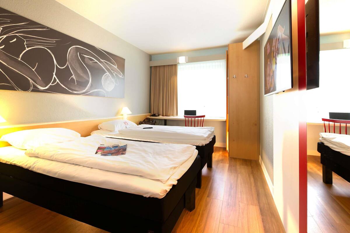 A hotel room with two beds, a desk, a chair, a TV, and a large abstract painting on the wall.