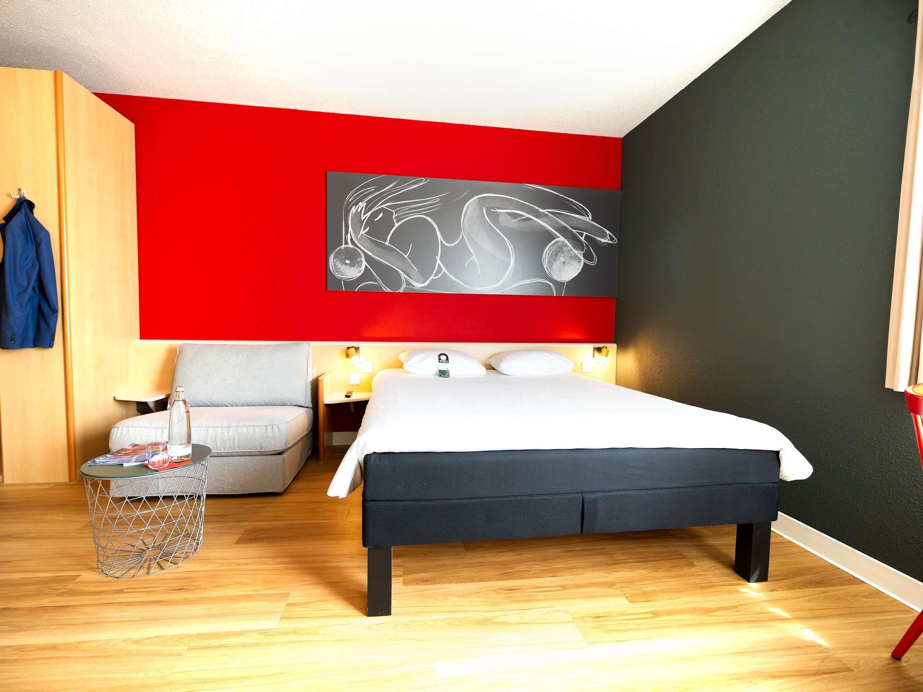 Una habitación de hotel moderna con una cama grande, un sillón gris, una mesita con una botella y una obra de arte en la pared.