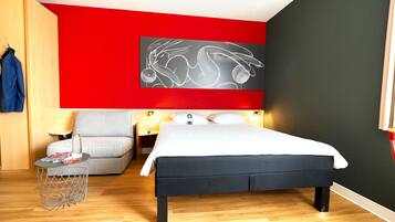 Una habitación de hotel moderna con una cama grande, un sillón gris, una mesita con una botella y una obra de arte en la pared.