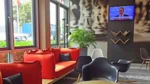 A modern waiting area with a red sofa, black chairs, a wooden table, and a television screen. - ibis Hotel Köln Messe (Cologne)