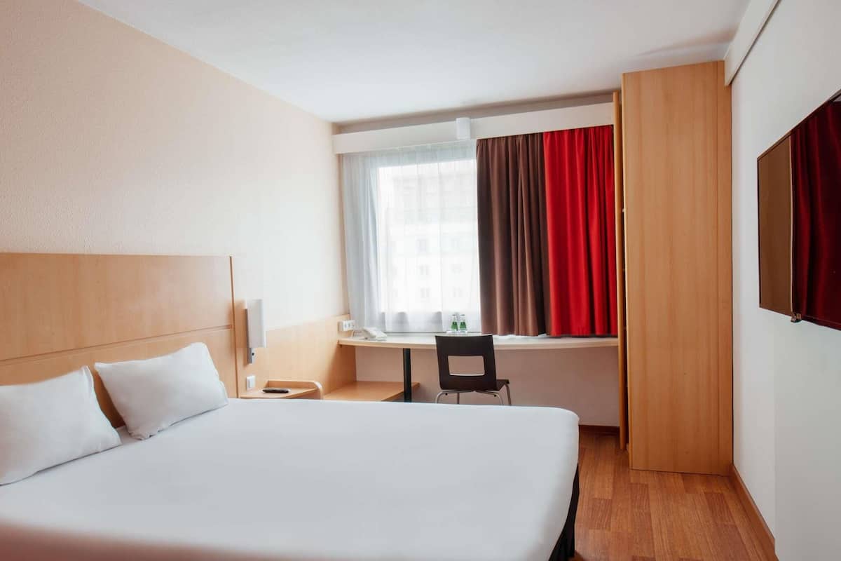 Ein Hotelzimmer mit einem großen Bett, einem Schreibtisch, einem Stuhl und einem Fenster mit Vorhängen.
