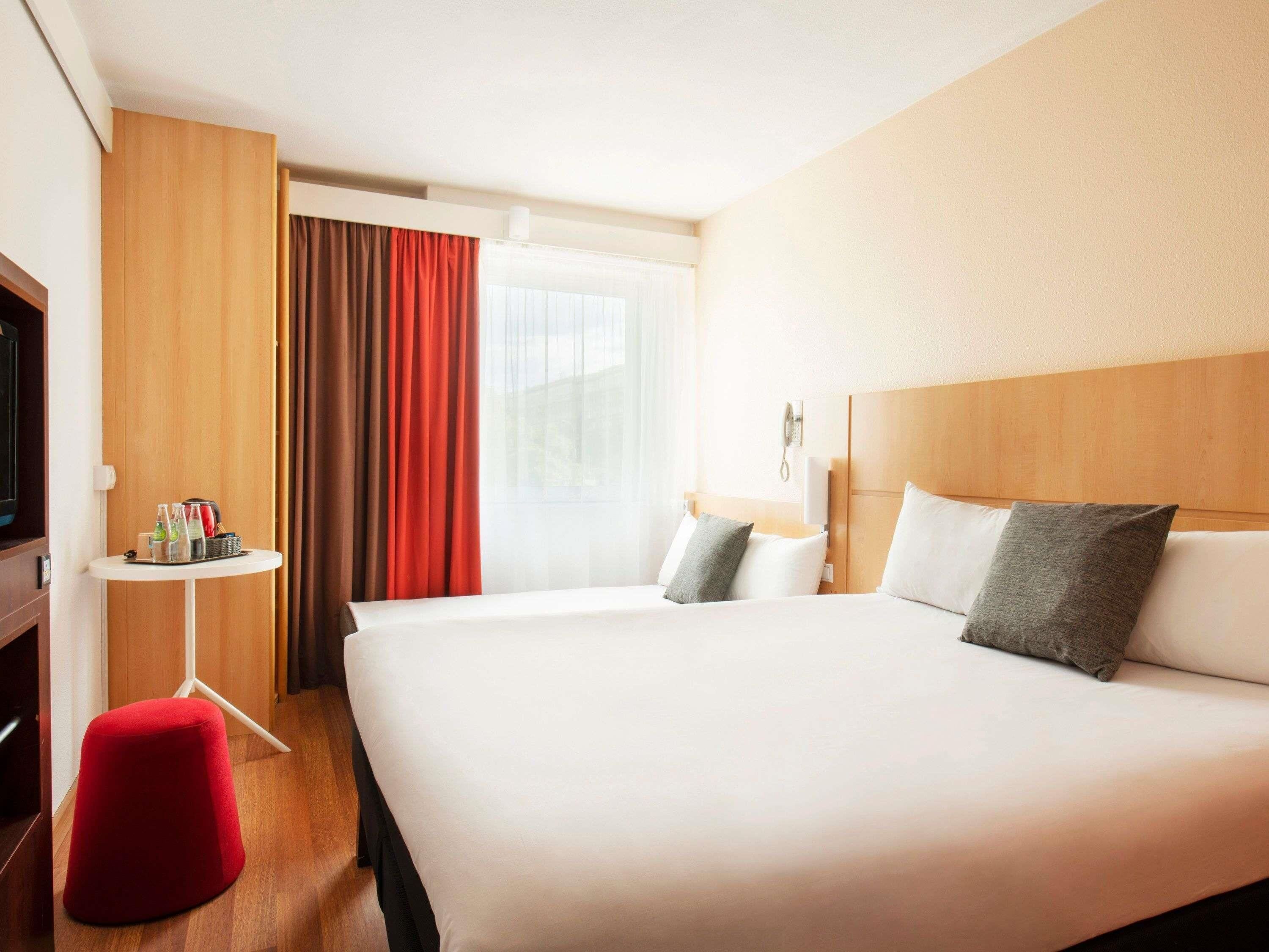 Habitación de hotel con una cama grande, una mesita con bebidas, un televisor y un taburete rojo.