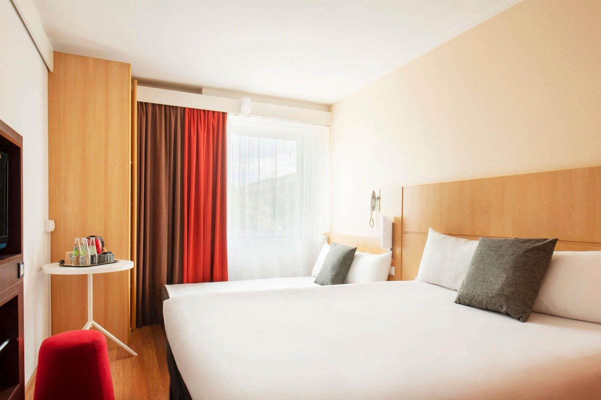 Habitación de hotel con una cama grande, una mesita con bebidas, un televisor y un taburete rojo.