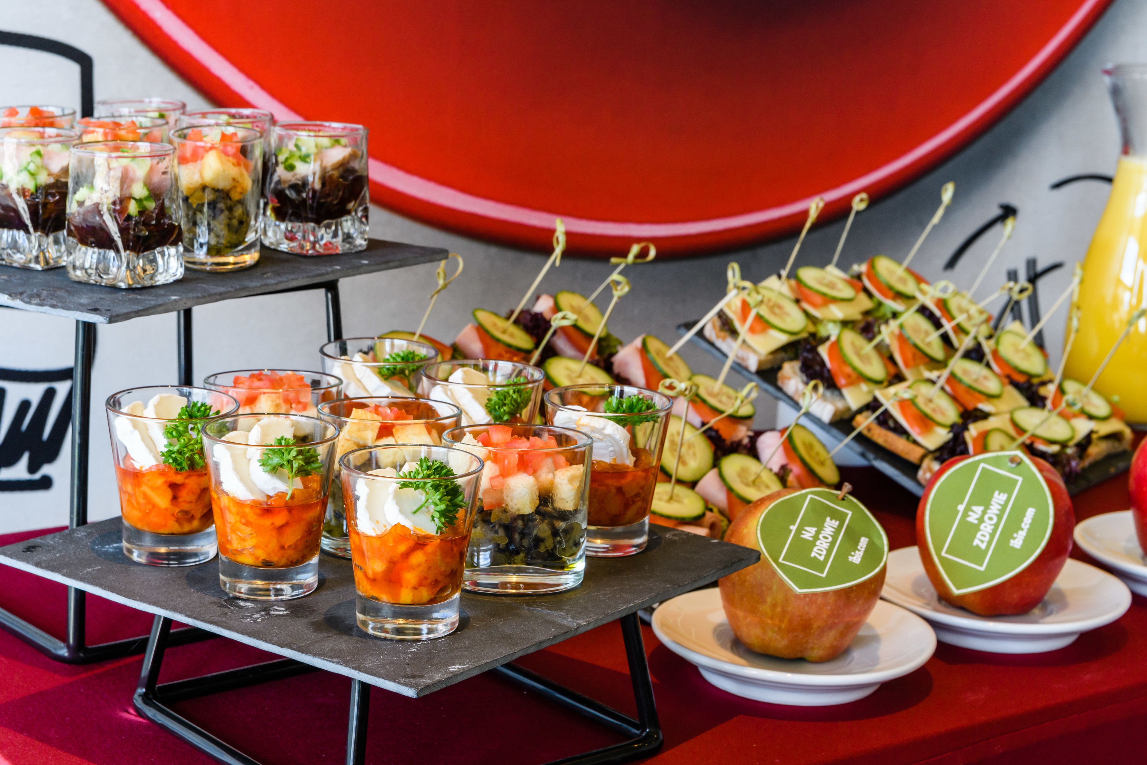 Diversos aperitivos, incluindo legumes e frutas espetadas, e pequenas taças com sobremesas em camadas, dispostos sobre uma mesa com fundo vermelho.