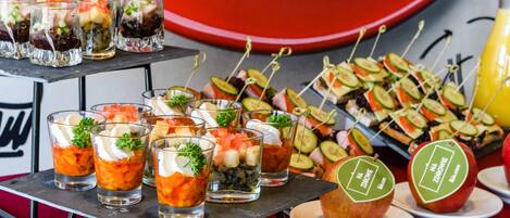 Verschiedene Vorspeisen, darunter Spieße mit Gemüse, Obst und kleine Gläser mit Schichtdesserts, auf einem Tisch mit rotem Hintergrund angerichtet.