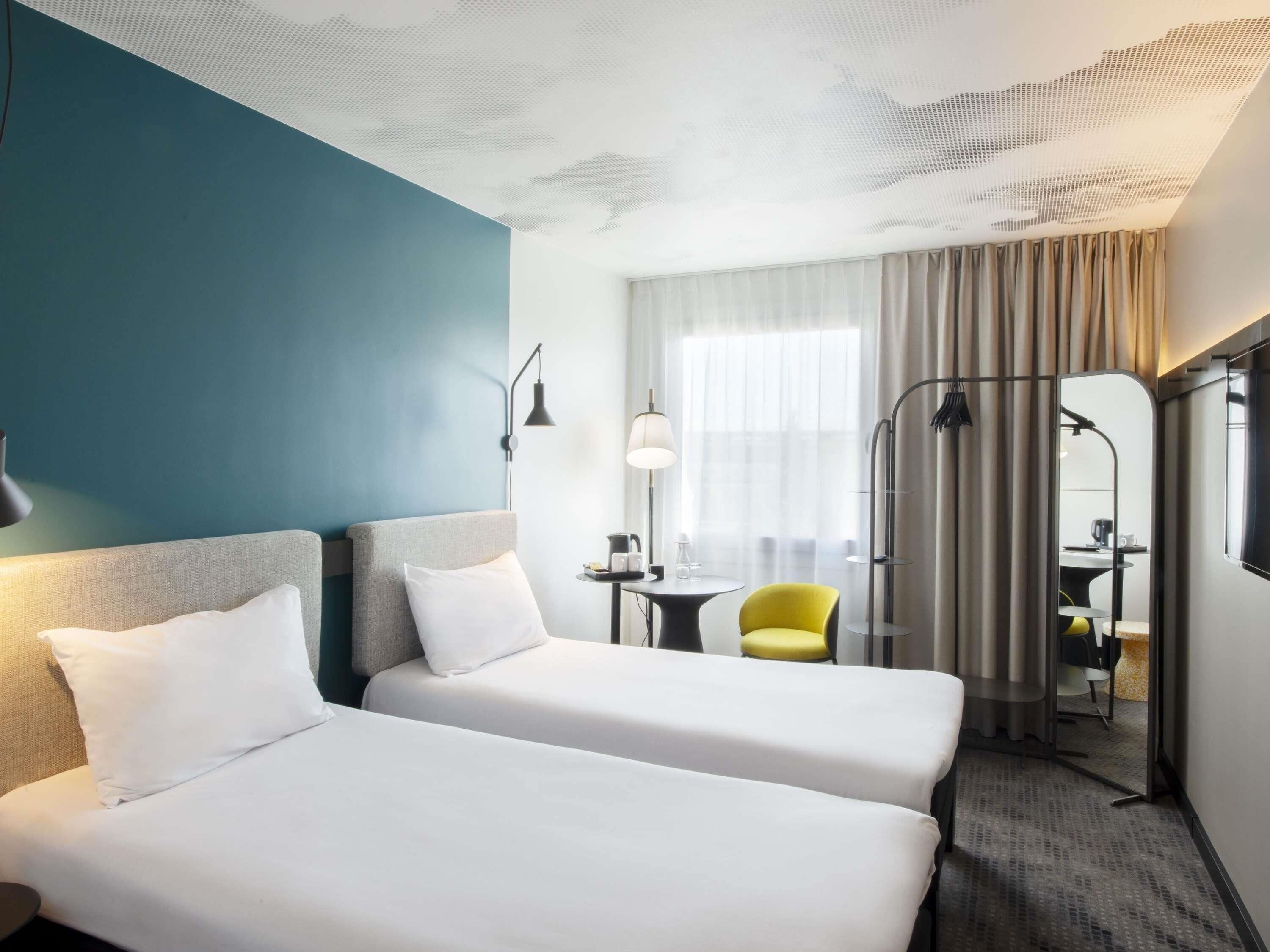 Habitación de hotel con dos camas, un escritorio, una silla y un ventanal con cortinas.