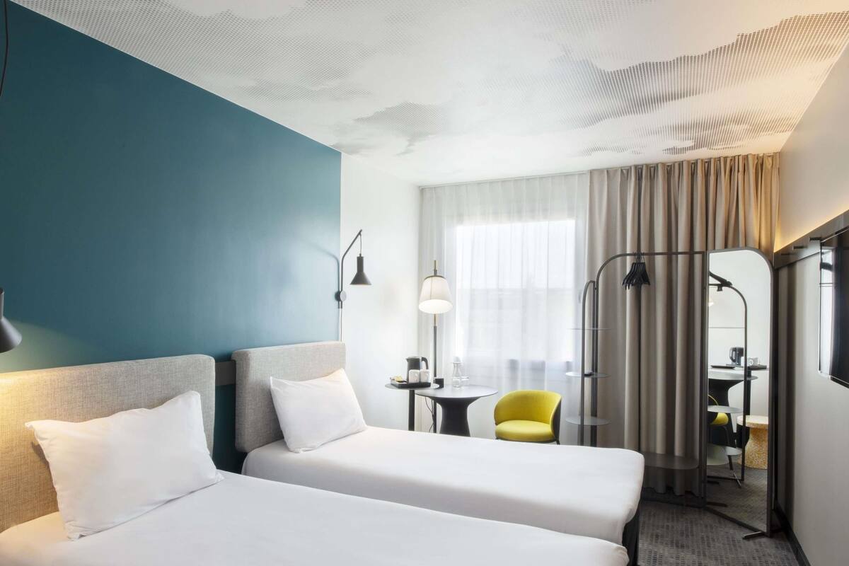 Habitación de hotel con dos camas, un escritorio, una silla y un ventanal con cortinas.
