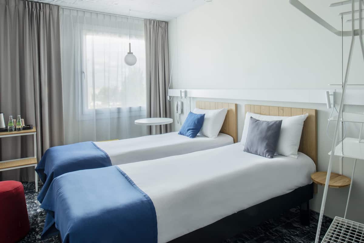 Habitación de hotel con dos camas, una mesita, una silla, una mesita de noche, una lámpara y un ventanal con cortinas.