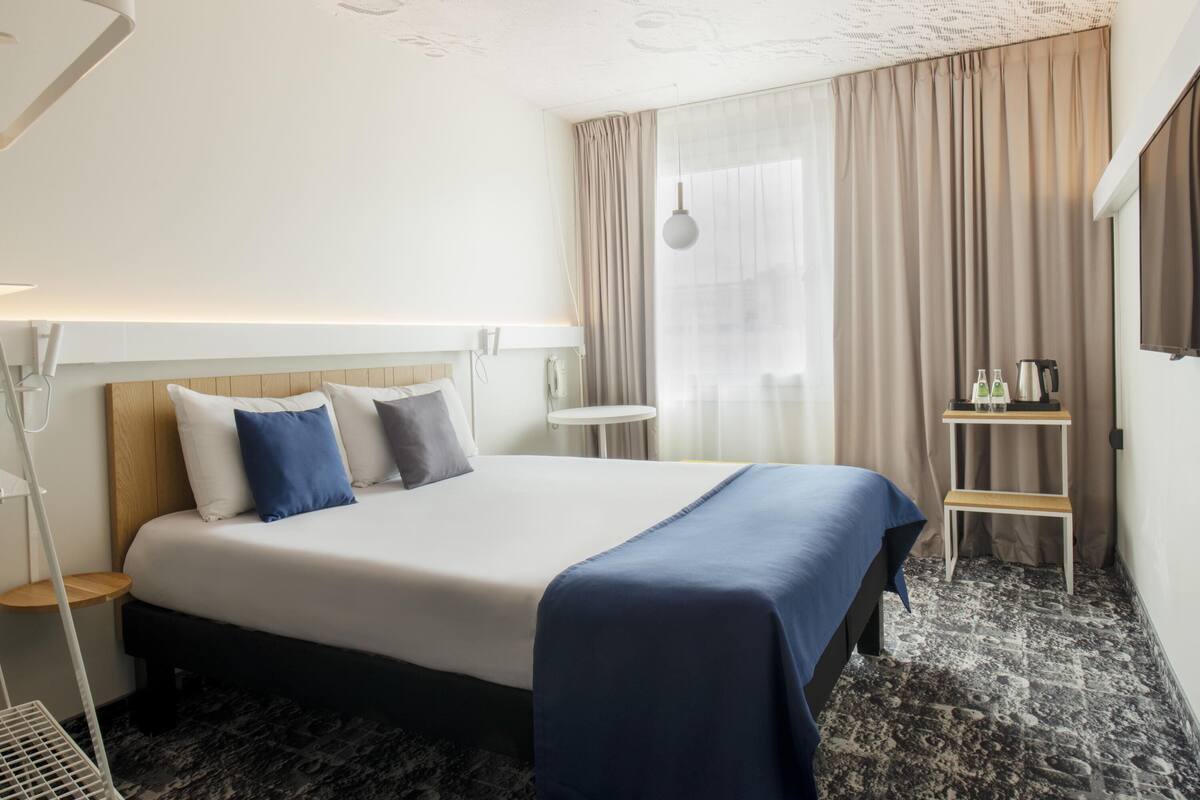 Habitación de hotel con cama, mesita de noche, lámpara y una pequeña mesa con botellas y un hervidor.