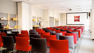 A conference room with rows of chairs, a projector screen displaying the ibis logo, and a presentation area with a desk and refreshments.