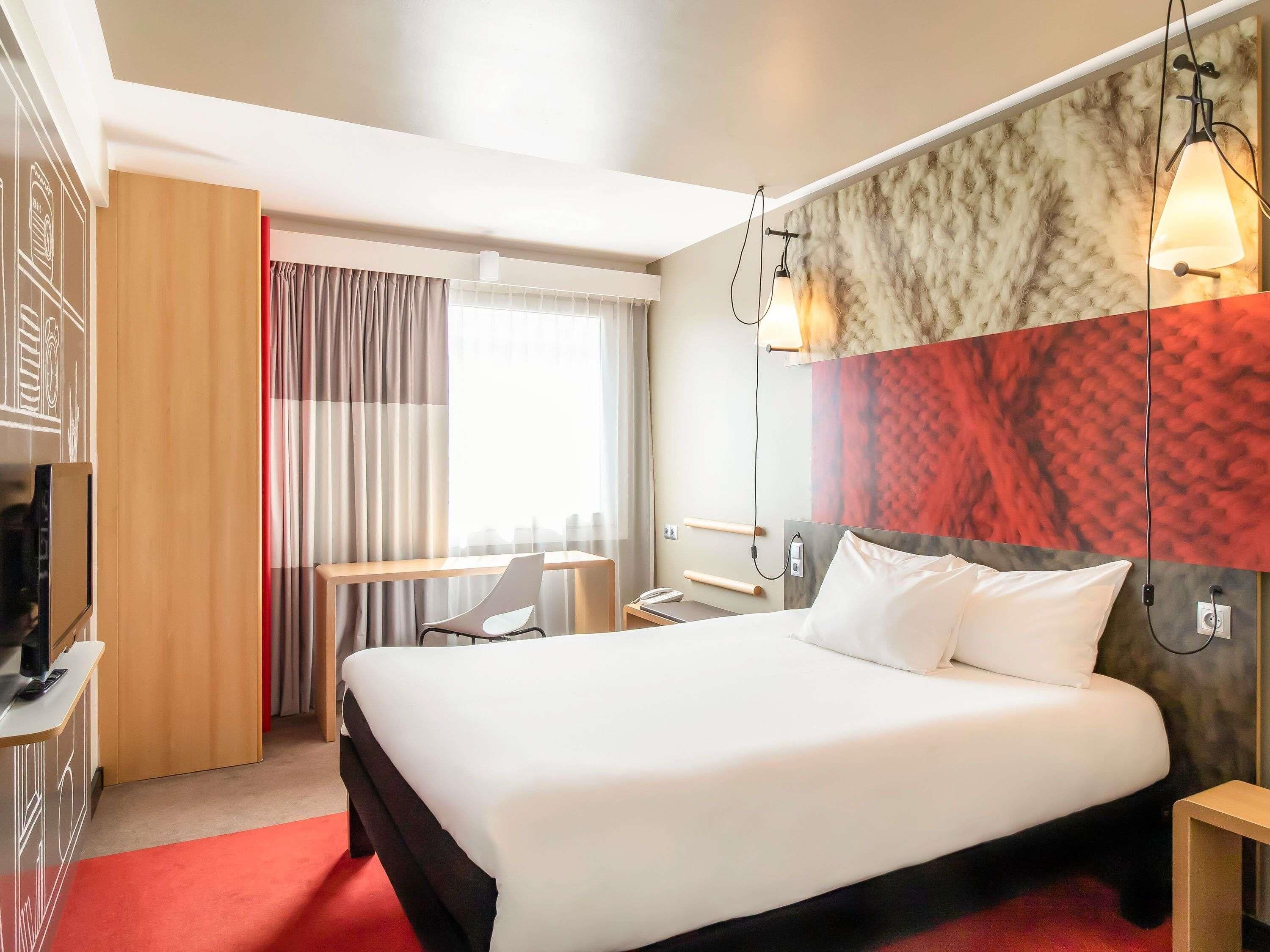 A hotel room with a large bed, a desk, a chair, a TV, and a window with curtains.