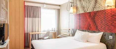 A hotel room with a large bed, a desk, a chair, a TV, and a window with curtains.