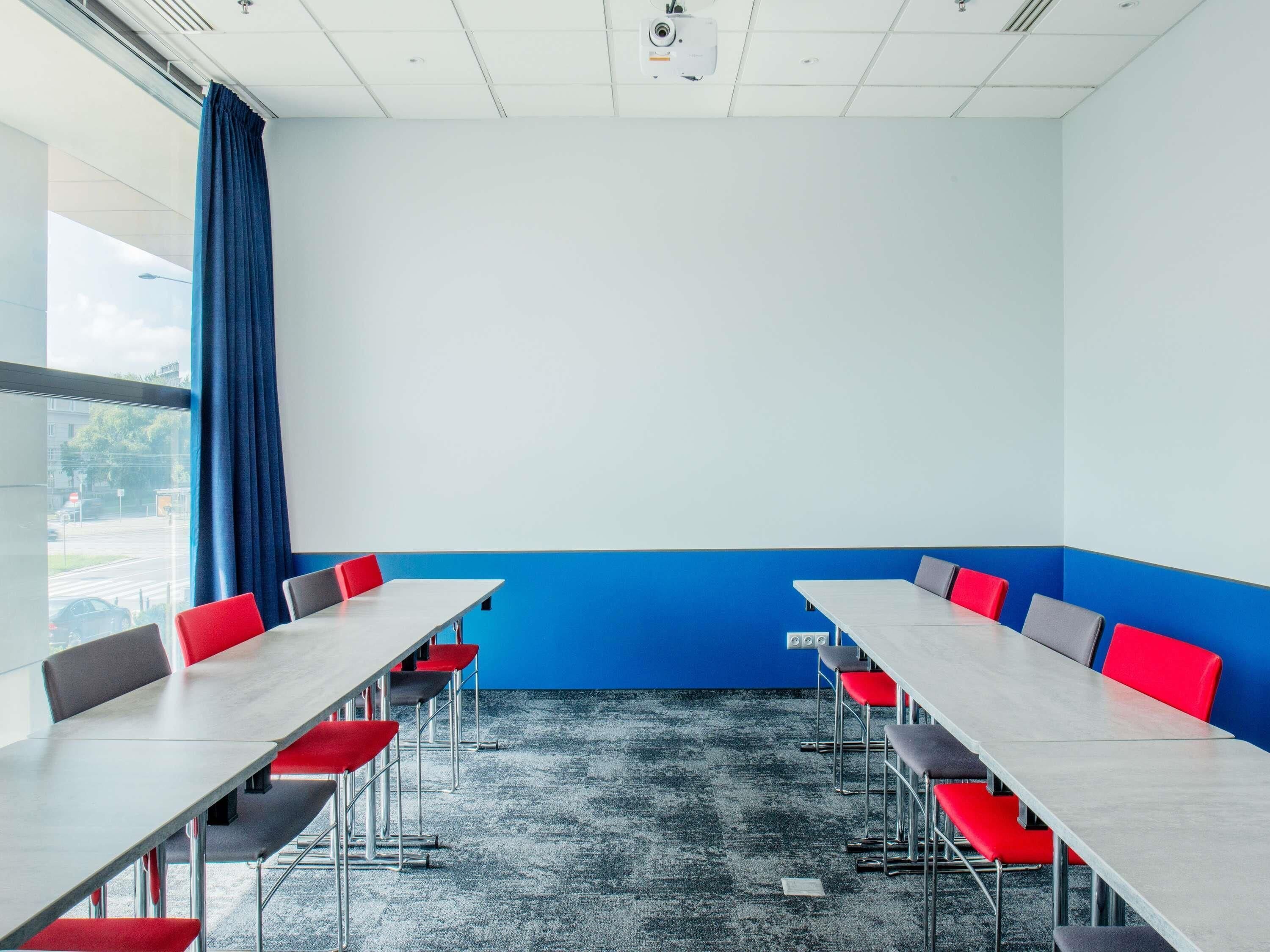 Un salón de conferencias con una larga mesa, sillas rojas y grises, una pared de acento azul y un ventanal grande.