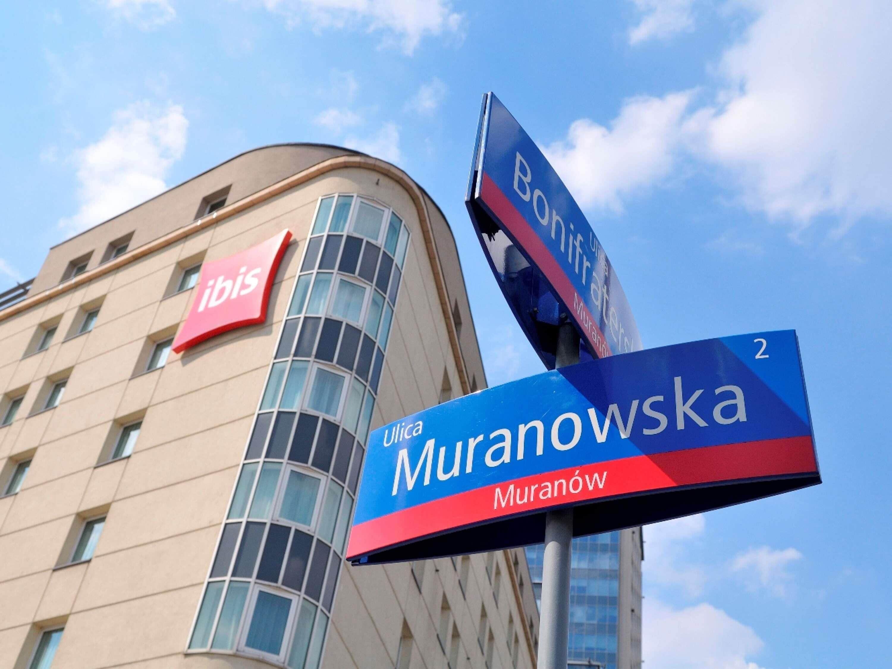 A hotel with a curved facade, a prominent red 'ibis' sign, and street signs for 'Muranowska' and 'Bonifacia'.