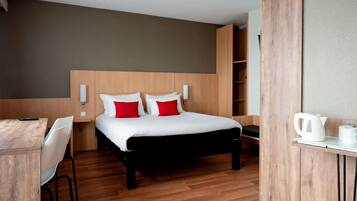 A modern hotel room with a wooden bed, white bedding, red pillows, a desk with a kettle, and a chair.
