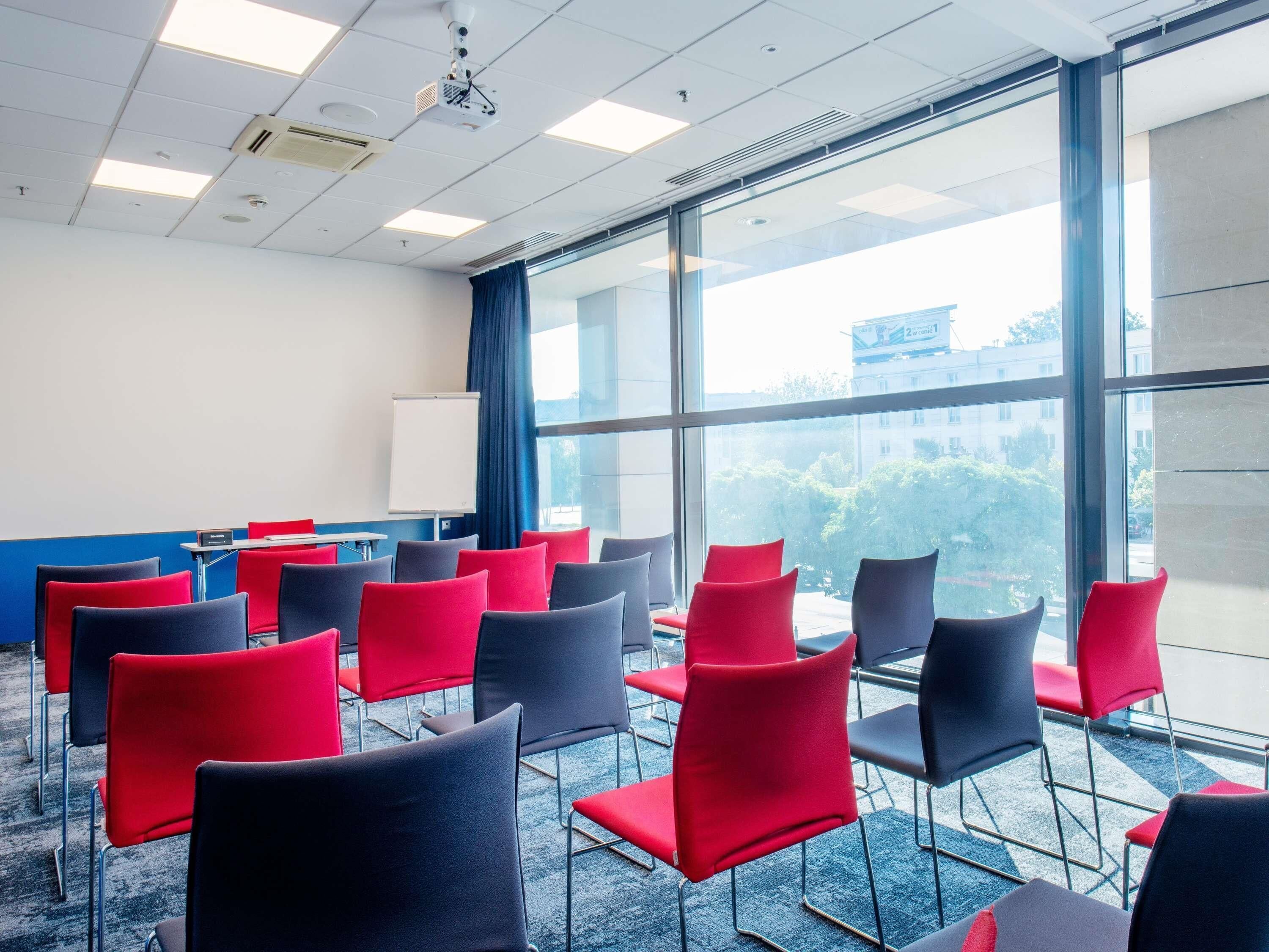 A conference room with rows of red and black chairs, a whiteboard, and a projector on the ceiling.