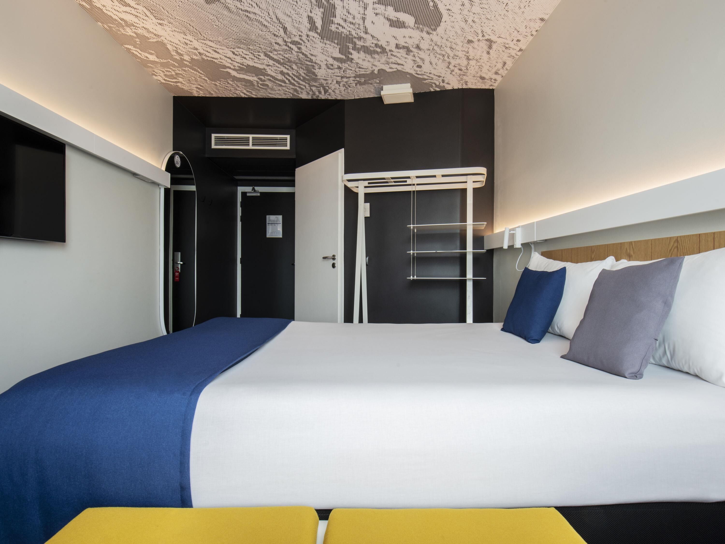 Una habitación de hotel moderna con una cama grande, un televisor de pantalla plana montado en la pared y un diseño de techo con una lámpara original.