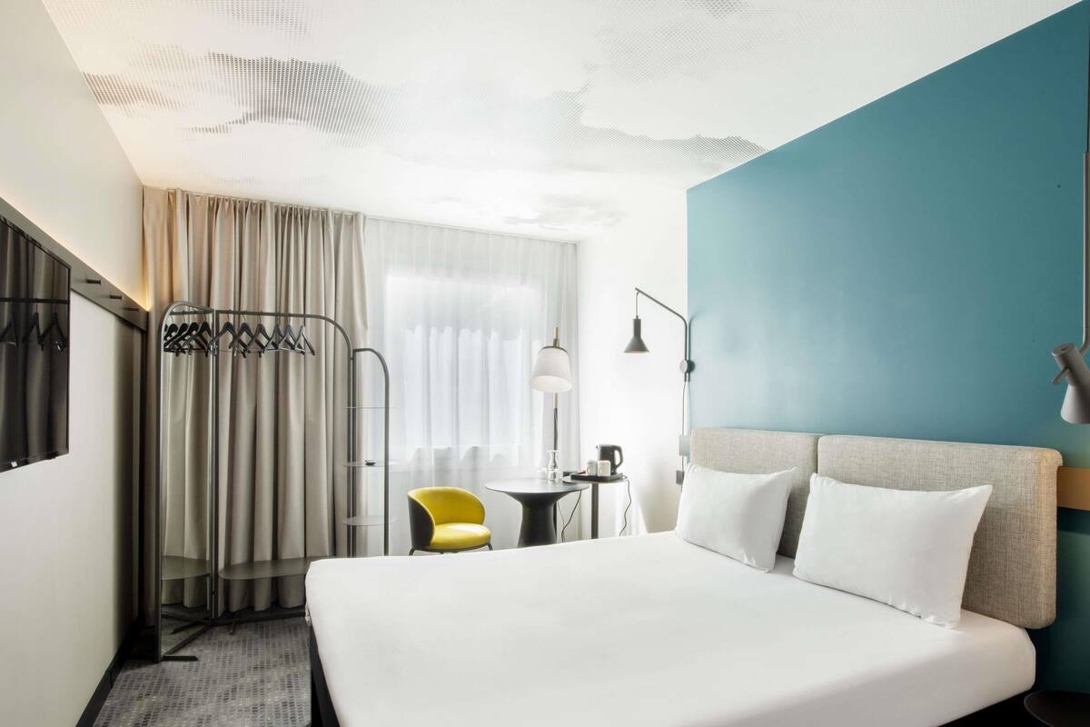 Una habitación de hotel con cama, escritorio, silla, armario y ventana con cortinas.