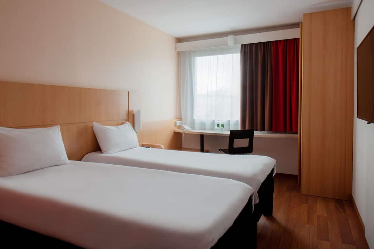 Habitación de hotel con dos camas, un escritorio, una silla y un ventanal con cortinas.