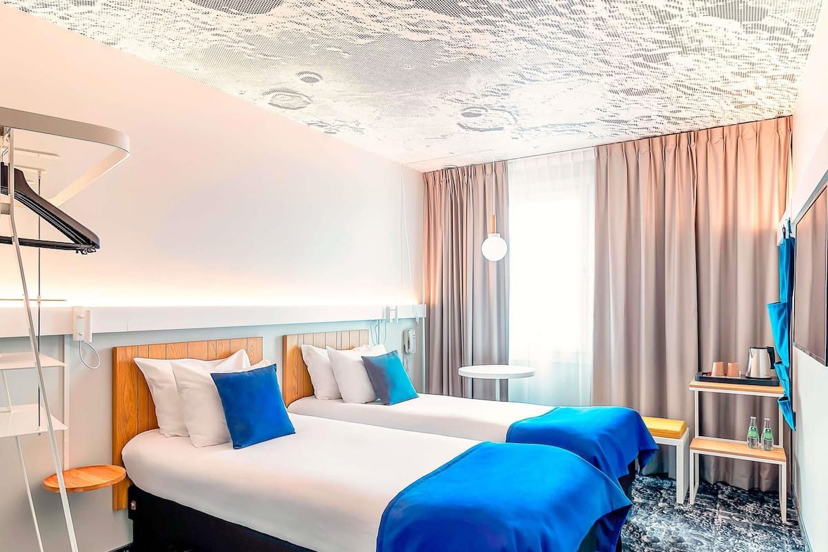 Ein Hotelzimmer mit zwei Betten, einem kleinen Tisch, einem Stuhl, einem Schreibtisch, einer Lampe und einem großen Fenster mit Vorhängen.