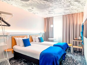 Ein Hotelzimmer mit zwei Betten, einem kleinen Tisch, einem Stuhl, einem Schreibtisch, einer Lampe und einem großen Fenster mit Vorhängen.