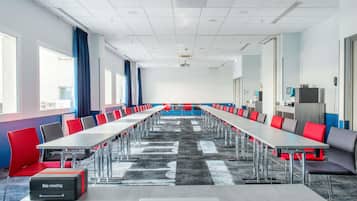 Une salle de conférence avec de longues tables, des chaises rouges et grises, un projecteur et une grande fenêtre.