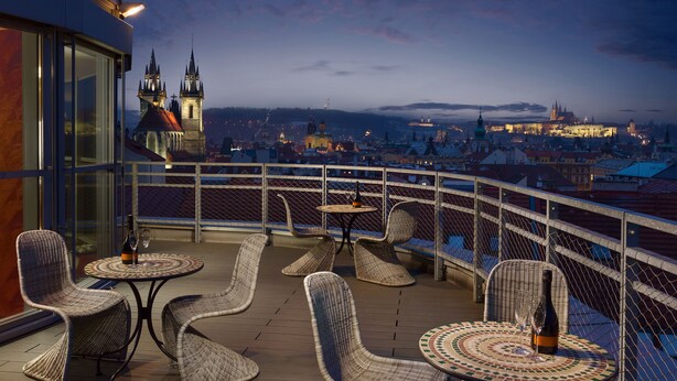 A terrace with wicker chairs and tables, overlooking a cityscape with historic buildings and a castle. - Hotel Astoria (Prague)