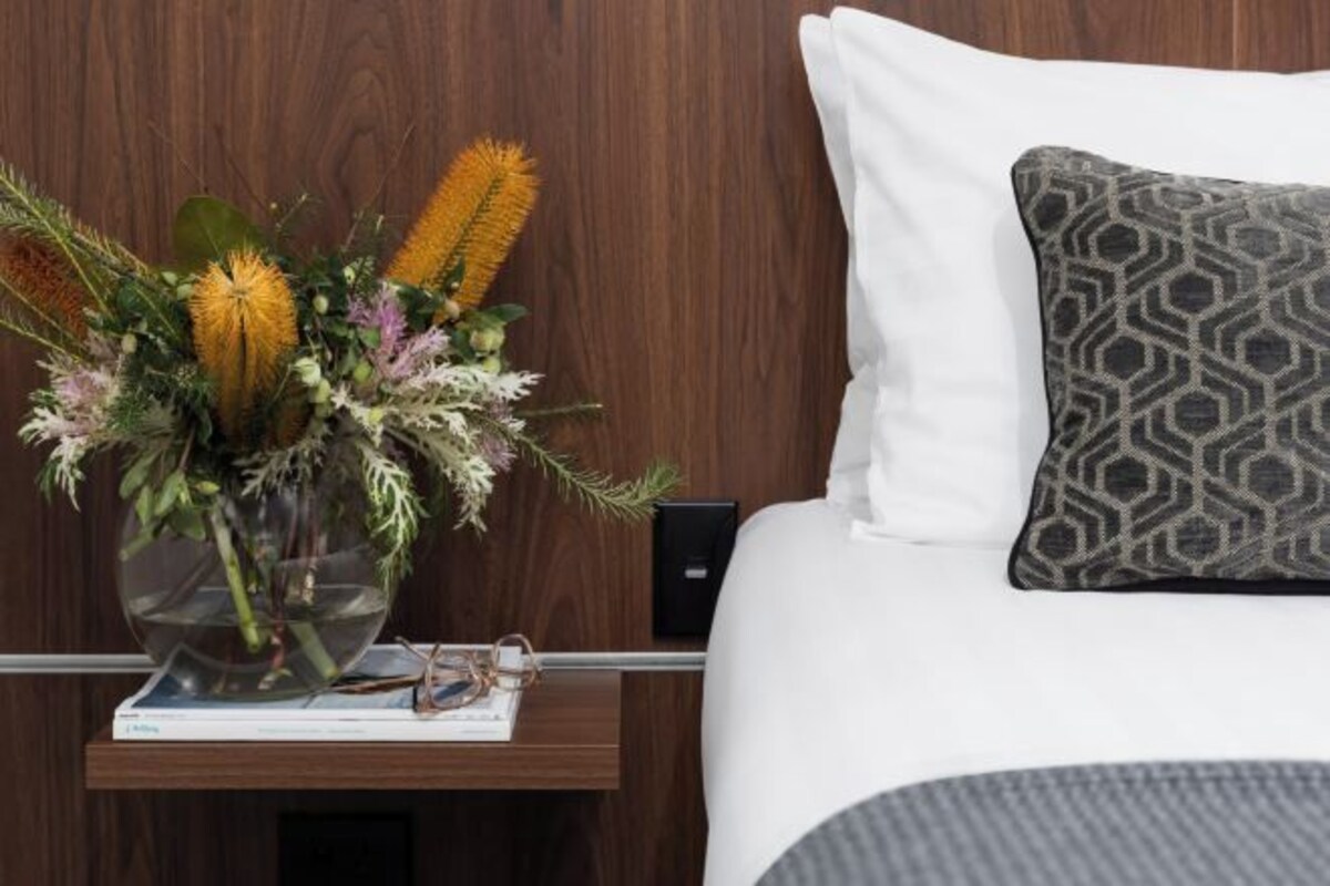A hotel room with a wooden headboard, a white bed with a patterned pillow, a bedside table with a vase of dried flowers, and a remote control.
