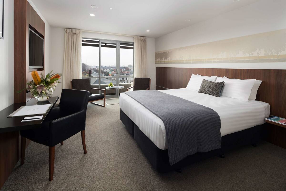 Habitación de hotel con una cama grande, un escritorio con silla, una mesita con un florero y vistas a la ciudad a través de amplios ventanales.