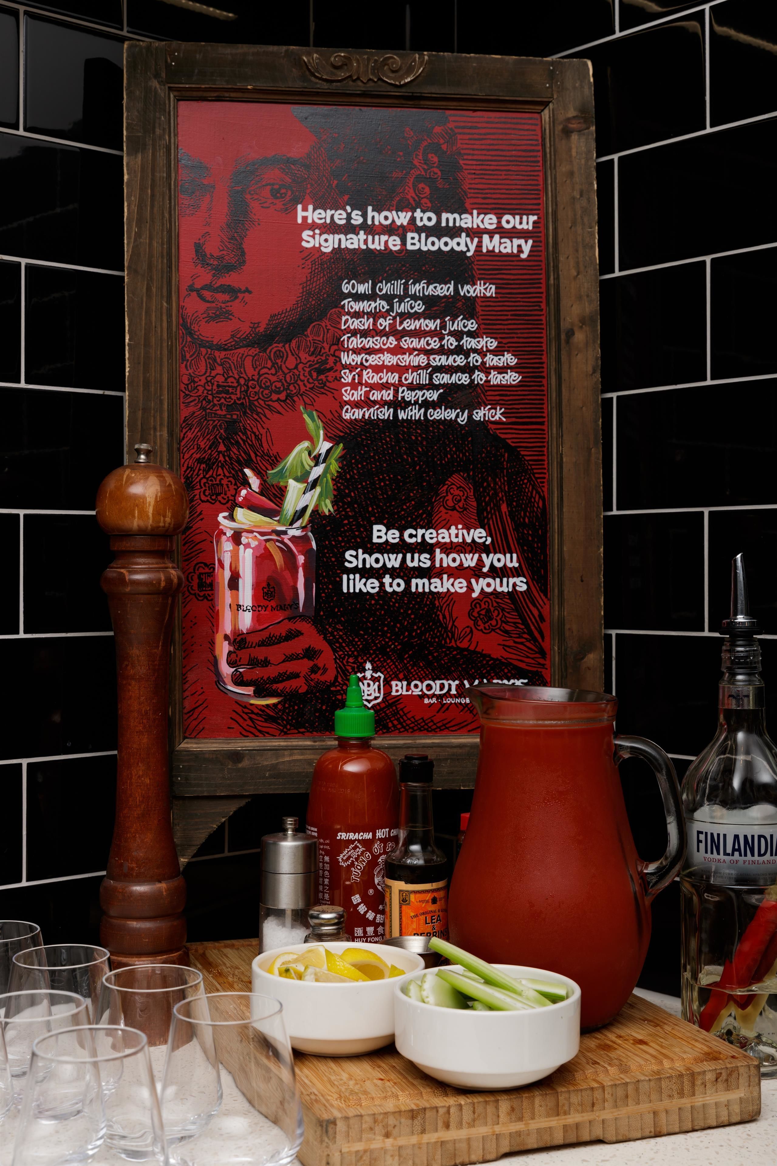 A kitchen counter with a recipe poster, a pepper mill, a bottle of hot sauce, a pitcher, and a bowl of lemons and celery.