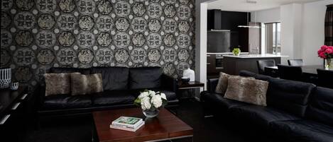 Una sala moderna con un sofá de cuero negro, una mesa de centro de madera con un libro y flores, y un papel tapiz estampado.