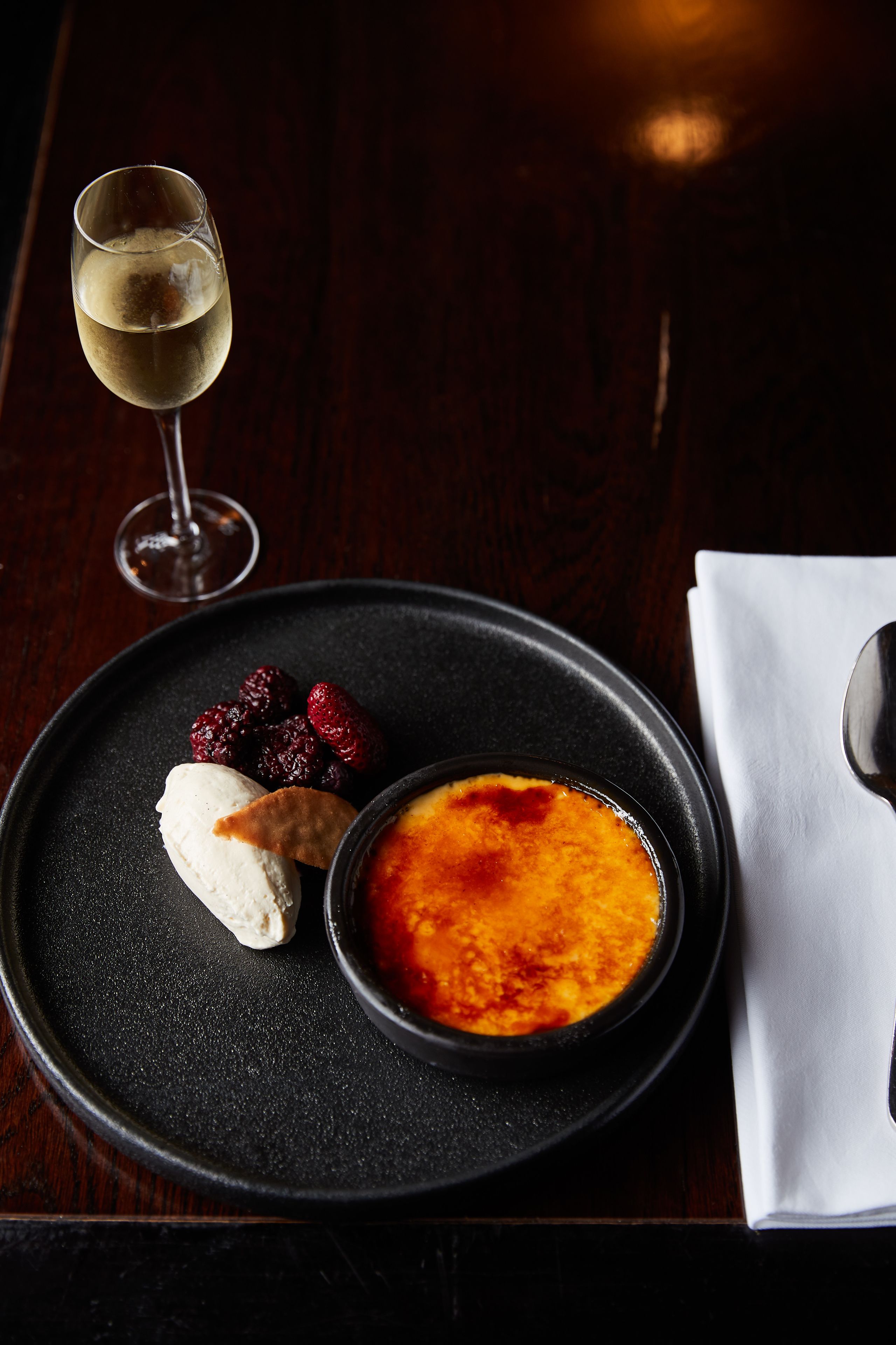 A dessert plate with a crème brûlée, berries, and a scoop of ice cream, accompanied by a glass of wine on a dark table.