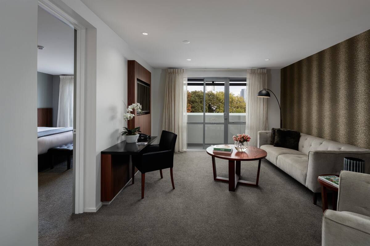 Habitación de hotel moderna con zona de comedor, sofá, mesa de centro y vistas a la ciudad a través de amplias ventanas.