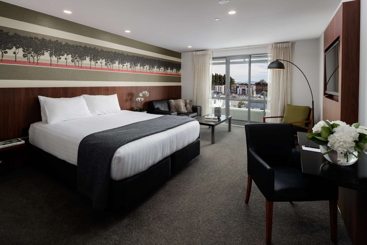 Habitación de hotel con una cama grande, un escritorio con silla, un sofá, una mesita de centro y vistas de la ciudad a través de amplias ventanas.