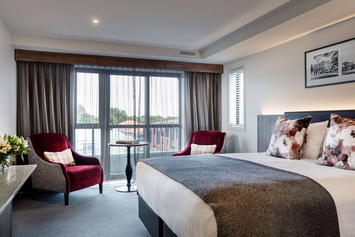 Habitación de hotel con una cama grande, dos sillones rojos, una mesita y vistas amplias a través de grandes ventanales.