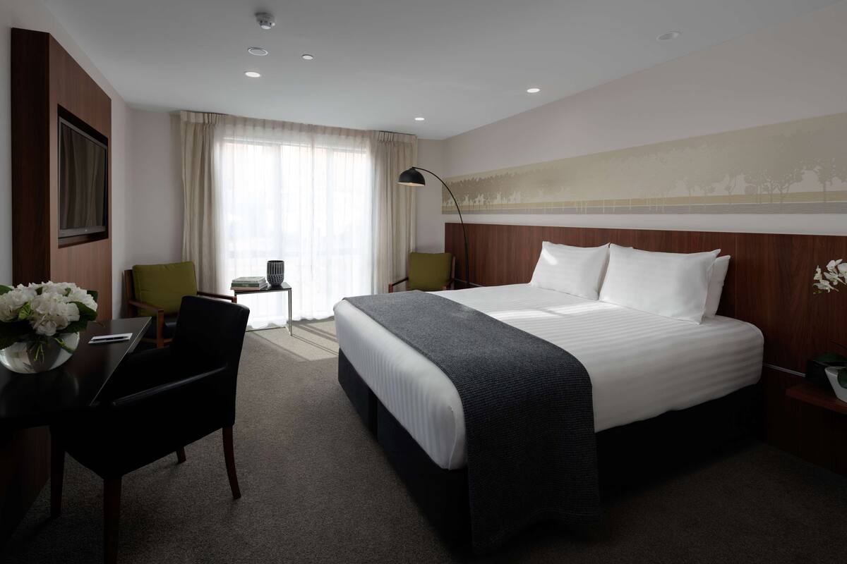 Habitación de hotel con una cama grande, un escritorio con silla, una lámpara y una ventana con cortinas.