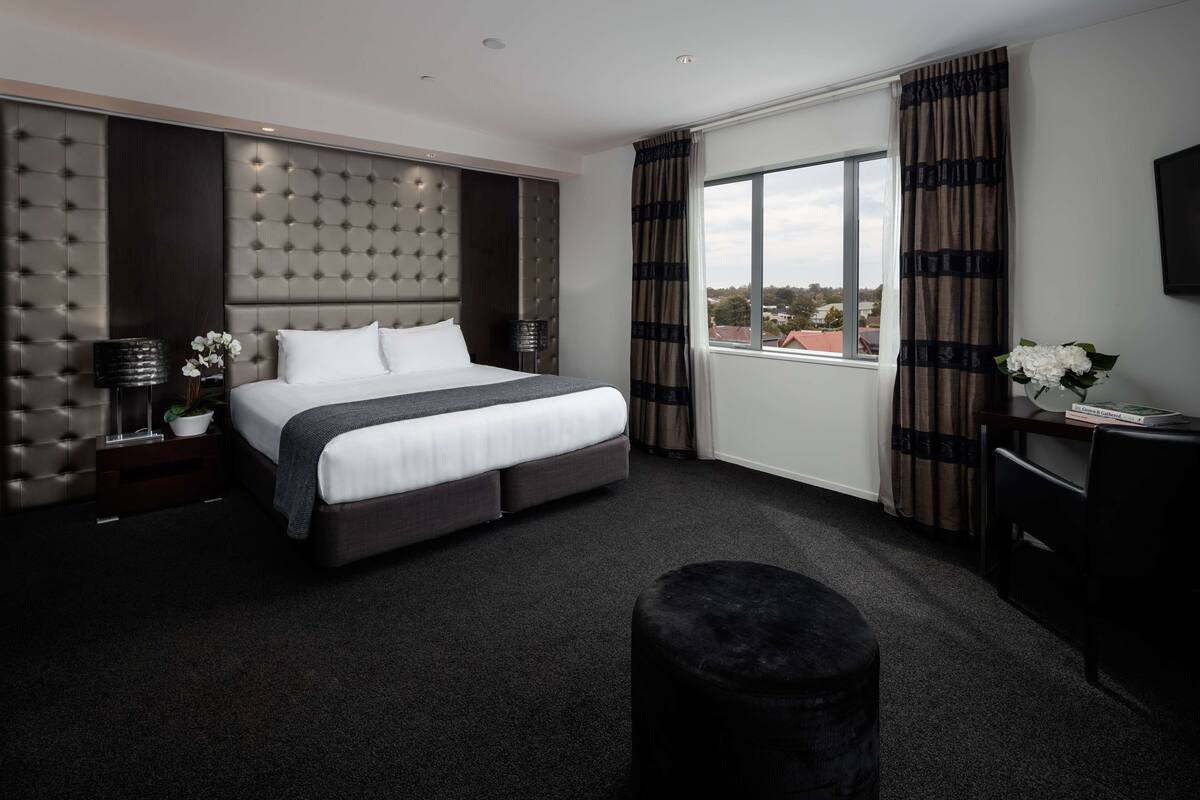 Una habitación de hotel moderna con una cama grande, alfombra oscura, un pequeño reposapiés, una mesita de noche con lámpara y vistas a la ciudad desde la ventana.