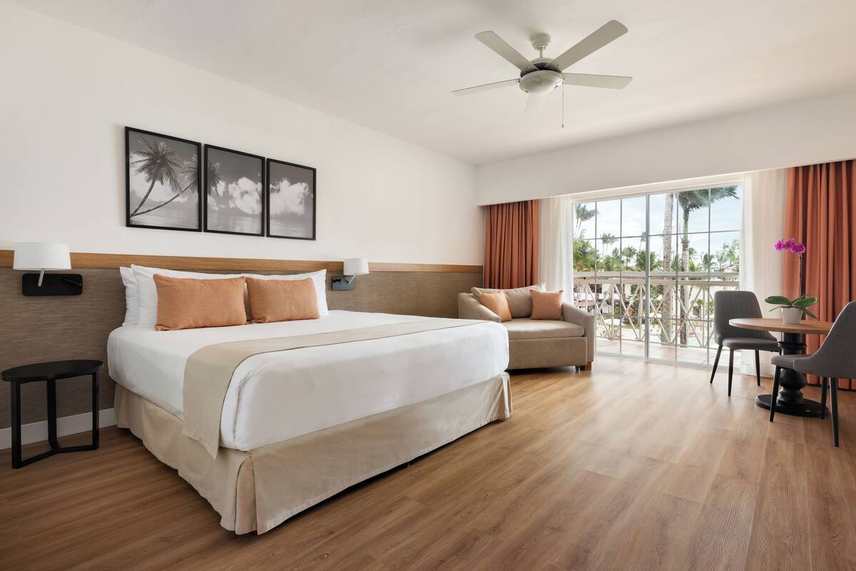 Habitación de hotel con una cama grande, un sofá, una mesa de comedor y un balcón con vistas.